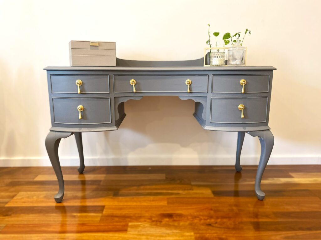 A stylish gray desk with golden knobs, featuring multiple drawers and elegant curved legs, adorned with a small box and decorative plants on top.